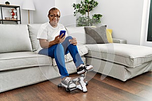 Senior african american woman using smartphone and doing sport using bike pedals machine at home