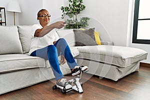Senior african american woman watching tv doing sport using bike pedals machine at home