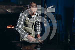 Senior an artisan blacksmith knocks with a hammer on iron to shape against the background of a burning forge