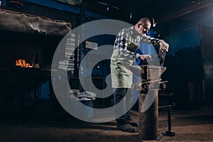 Senior an artisan blacksmith knocks with a hammer on iron to shape against the background of a burning forge