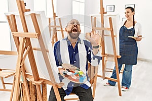 Senior artist man at art studio crazy and mad shouting and yelling with aggressive expression and arms raised