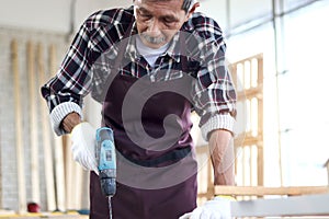 Senior Asian carpenter work in woodwork workshop, old citizen craftsman working with hand drill in the joinery, cheerful elderly