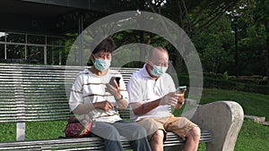 Senior Asian couple with face mask sitting on a bench and using their phones.
