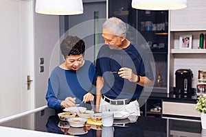 Senior Asian couple grandparents cooking together while woman is feeding food to man at the kitchen. Long lasting relationship