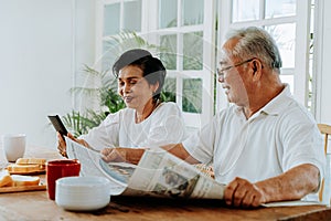 Senior Asian couple having breakfast together in dinner room. 70s retired elderly man reading newspaper while woman
