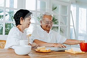 Senior Asian couple having breakfast together in dinner room. 70s retired elderly man and woman reading newspaper