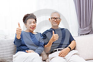 Senior Asian couple looking at camera and giving thumbs up while sitting on sofa at home.