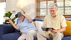 Senior asian couple playing virtual reality headset and using digital tablet in home living room with happiness emotion,