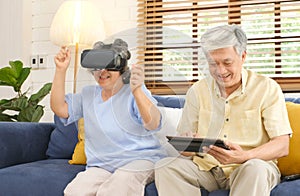 Senior asian couple playing virtual reality headset and using digital tablet in home living room with happiness emotion,