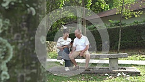 Senior Asian couple sitting on bench in a park, reading and surfing phone together.