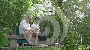 Senior Asian couple sitting on bench in a park, reading their mobile phones.