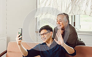 Senior asian father and adult son using smart phone talking on video call in living room