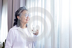 Senior Asian lady drinking glass of milk while standing by the window with copy space for calcium boosting and healthy dairy