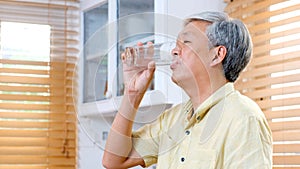 Senior asian man drinking water while standing by window in kitchen background, elderly retirement people and healthy lifestyles