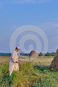 An senior asian man in an embroidered skullcap and white traditional Clothes mows hand-scythe grass in a hayfield