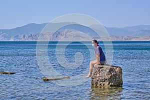 Senior asian man in face mask sits alone on a stone in a sea bay