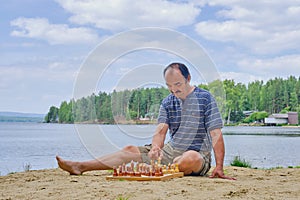 Senior asian man making a move in a game of chess and sitting on the beach near the lake