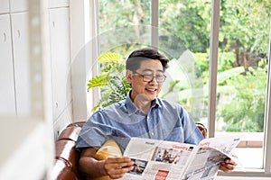 Senior Asian man reading newspaper mock up at the sofa couch inside of the house in the morning. Happy retirement with relaxation