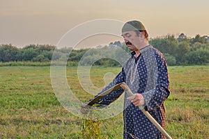 An senior asian man in a skullcap and traditional clothes cleans a hand scythe with grass in a hayfield