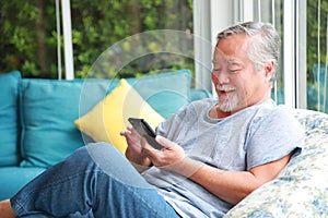 Senior Asian man using mobile phone while sitting in relaxation on the sofa couch in his home