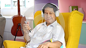 Senior asian man using smartphone   listening music with headphones, sitting on sofa in living room at home, Old male relaxation photo