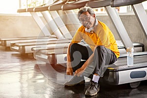 Senior asian sport man tying shoelaces on treadmill in fitness gym ready walking with headphone and water bottle . elder male