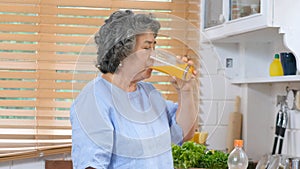 Senior asian woman drinking orange juice while standing by window in kitchen background, Old people retirement and healthy