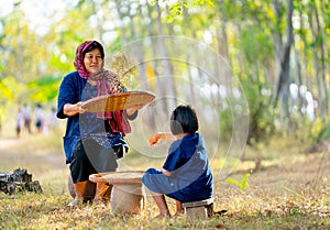Senior Asian woman with traditional clothes winnow rice using basketry and little girl stay beside and also work with rice