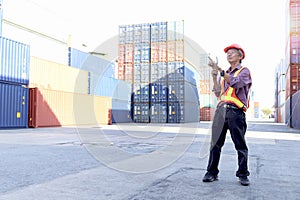 Senior Asian worker engineer wearing safety vest and helmet holding radio walkies talkie, standing and pointing away at logistic