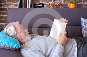 Senior attractive man lying down on sofa reading a book.  Old retired man relaxing at home.  Brick wall background