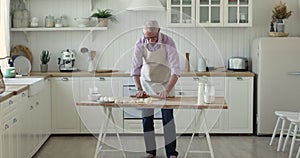 Senior baker man in apron rolling dough on floury table