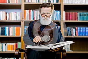 Senior bearded man in glasses, academic professor or teacher, sitting and reading an old book in the library, holding