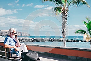 Senior bearded man holding smartphone sitting outdoors on bench face the sea enjoying sunny day, travel, vacation, retirement