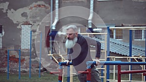 Senior bearded man with strong muscular arms exercising outdoors, doing pull ups, elderly male performing pulling exercise on hori