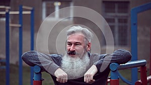 Senior bearded man with strong muscular arms exercising outdoors, doing pull ups, elderly male performing pulling exercise on hori