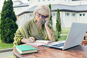 Senior beautiful business woman sitting on a chair and making notes in a notebook, call by phone and laptop in the yard