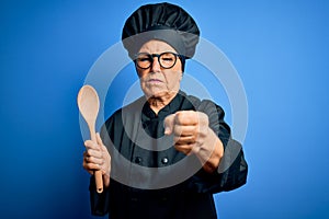 Senior beautiful grey-haired chef woman wearing cooker uniform and hat holding wooden spoon annoyed and frustrated shouting with
