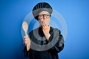 Senior beautiful grey-haired chef woman wearing cooker uniform and hat holding wooden spoon cover mouth with hand shocked with