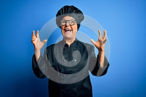 Senior beautiful grey-haired chef woman wearing cooker uniform and hat over blue background crazy and mad shouting and yelling