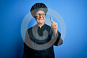 Senior beautiful grey-haired chef woman wearing cooker uniform and hat over blue background showing and pointing up with finger