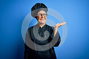 Senior beautiful grey-haired chef woman wearing cooker uniform and hat over blue background smiling cheerful presenting and