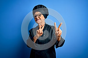 Senior beautiful grey-haired chef woman wearing cooker uniform and hat over blue background smiling looking to the camera showing