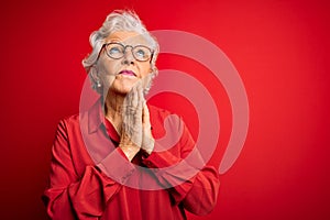 Senior beautiful grey-haired woman wearing casual shirt and glasses over red background begging and praying with hands together