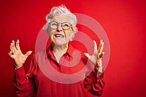 Senior beautiful grey-haired woman wearing casual shirt and glasses over red background crazy and mad shouting and yelling with