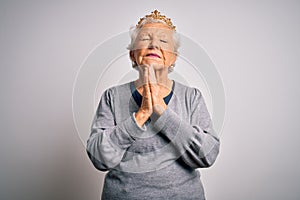 Senior beautiful grey-haired woman wearing golden queen crown over white background begging and praying with hands together with