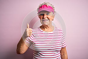 Senior beautiful sporty woman wearing sport cap standing over isolated pink background doing happy thumbs up gesture with hand