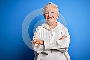 Senior beautiful sporty woman wearing white sweatshirt over isolated blue background happy face smiling with crossed arms looking