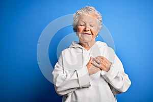 Senior beautiful sporty woman wearing white sweatshirt over isolated blue background smiling with hands on chest with closed eyes