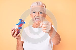 Senior beautiful woman with blue eyes and grey hair holding heart organ annoyed and frustrated shouting with anger, yelling crazy