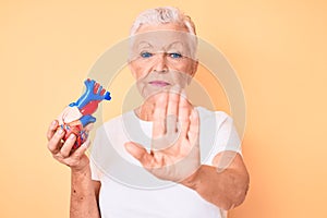 Senior beautiful woman with blue eyes and grey hair holding heart organ with open hand doing stop sign with serious and confident